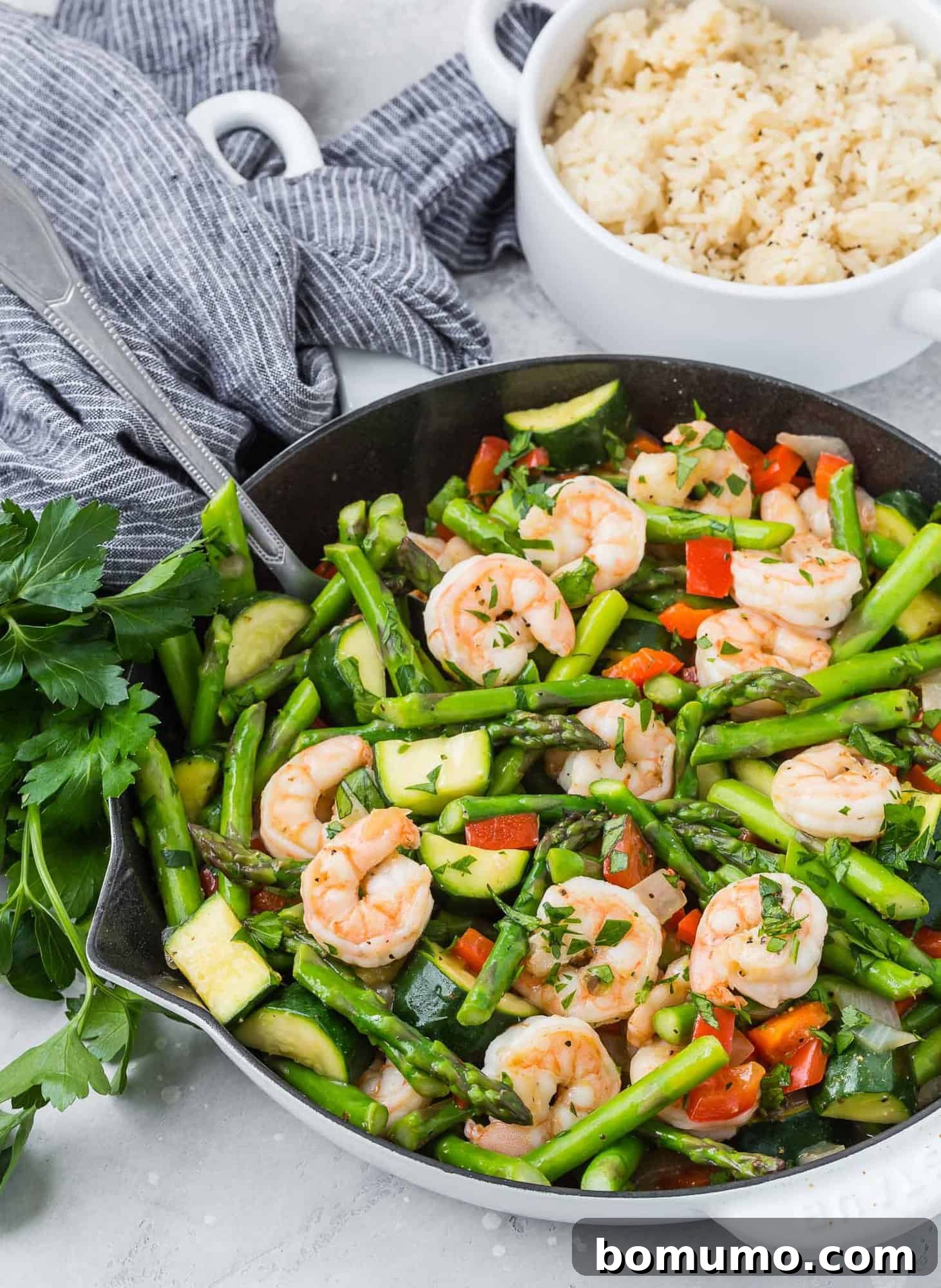 Shrimp stir fry in a white skillet, with fluffy cooked rice served in a bowl in the background.