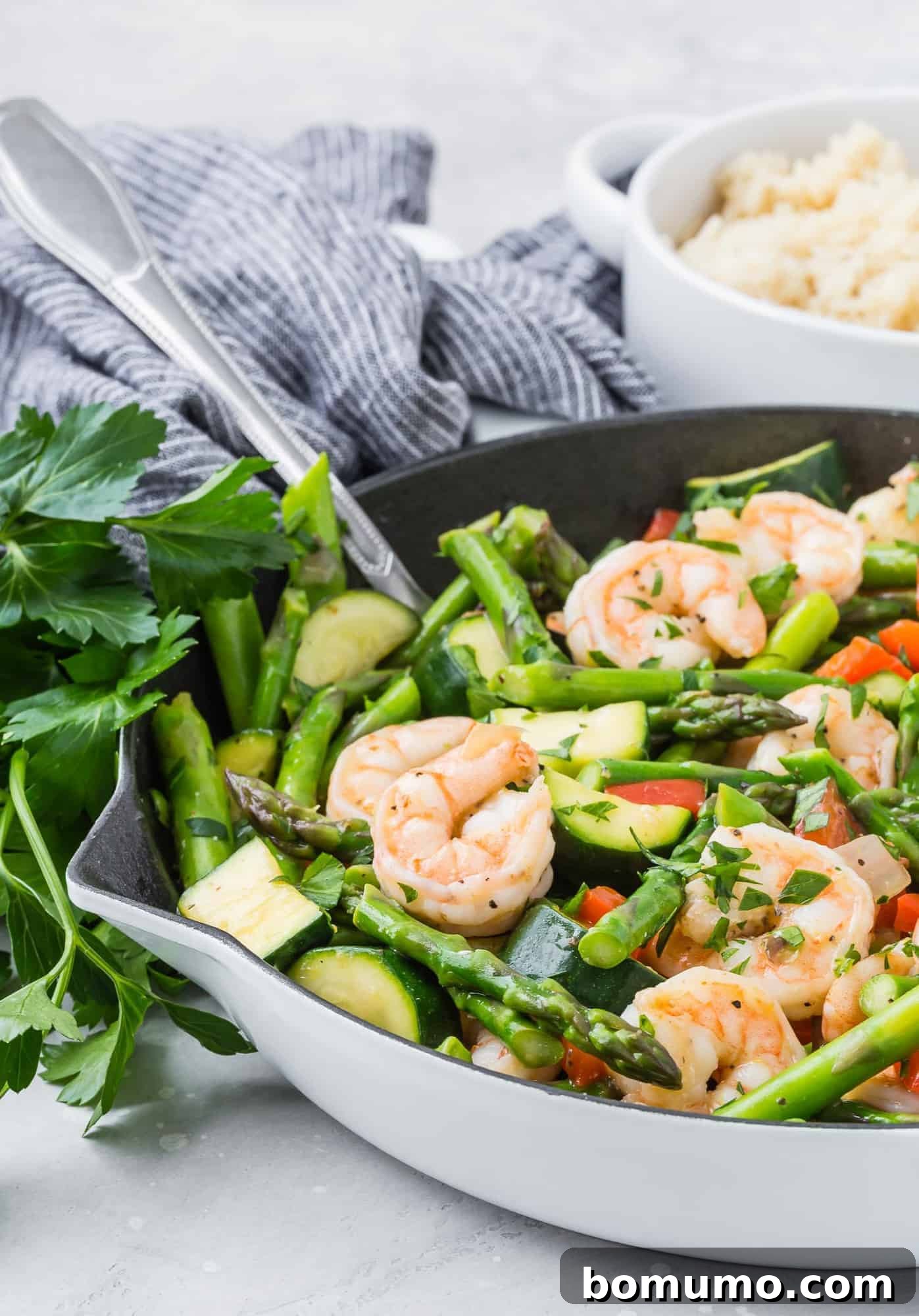 Stir fry with succulent shrimp and crisp asparagus in a white skillet, accompanied by a silver spoon.
