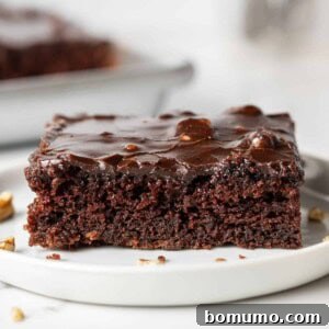 Square piece of chocolate cake with thin frosting, on a white plate.