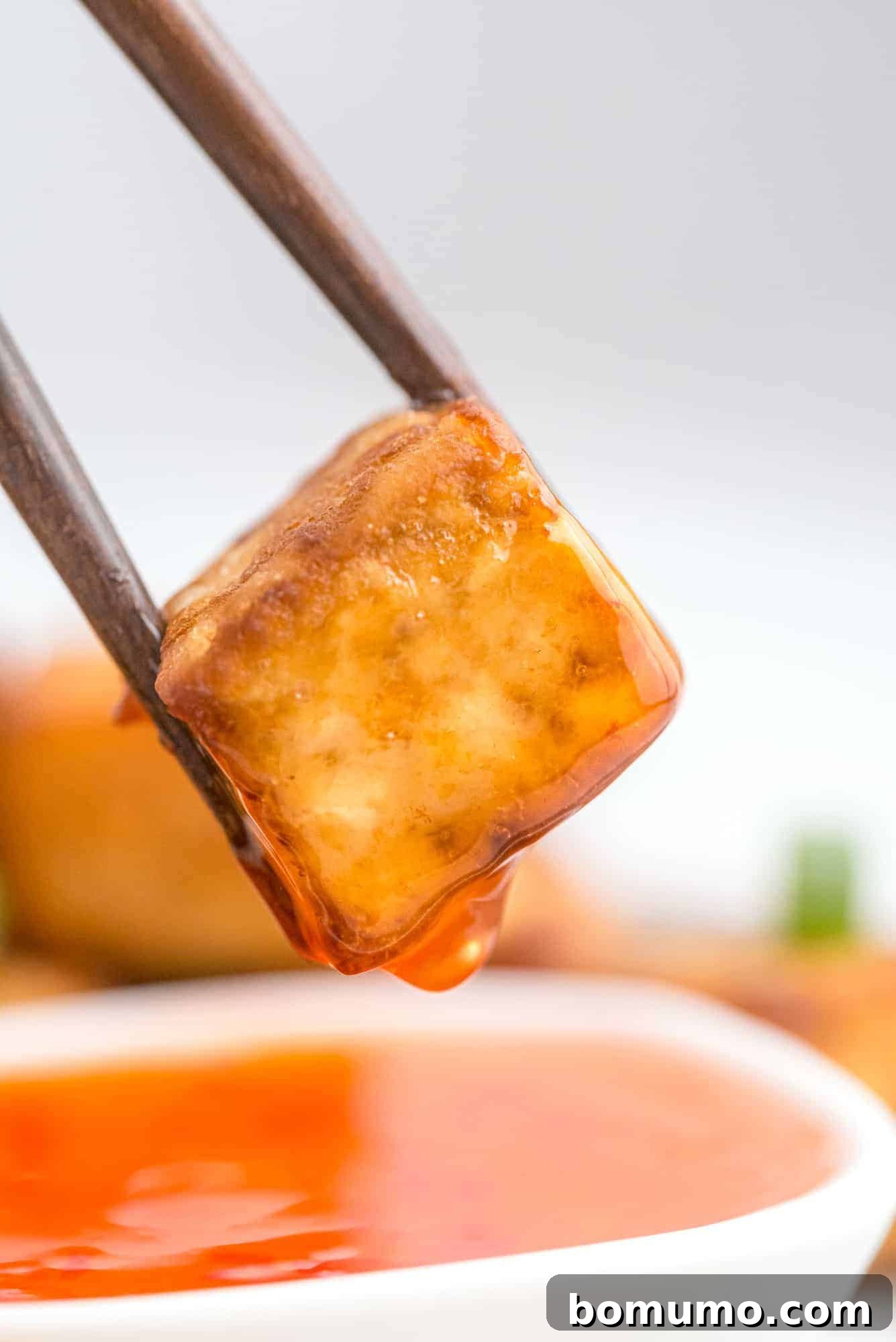 Tofu cube between chopsticks, with a drip of sweet chili sauce dripping off it.