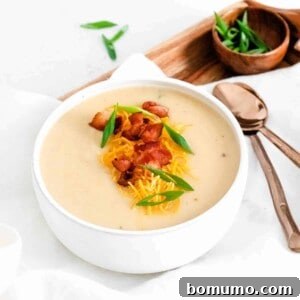 Cauliflower soup in a white bowl, topped with green onions, cheddar, and bacon. Spoons also visible.