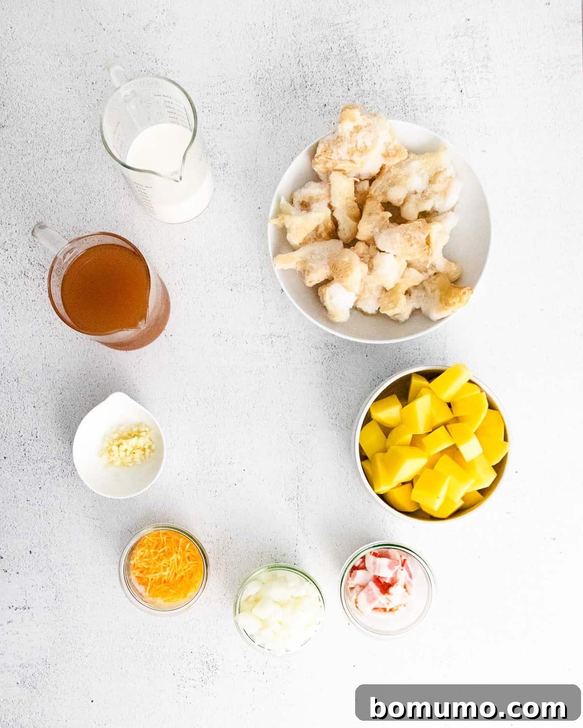 Overhead view of ingredients in bowls: potato, cauliflower, broth, milk, cheese, bacon.