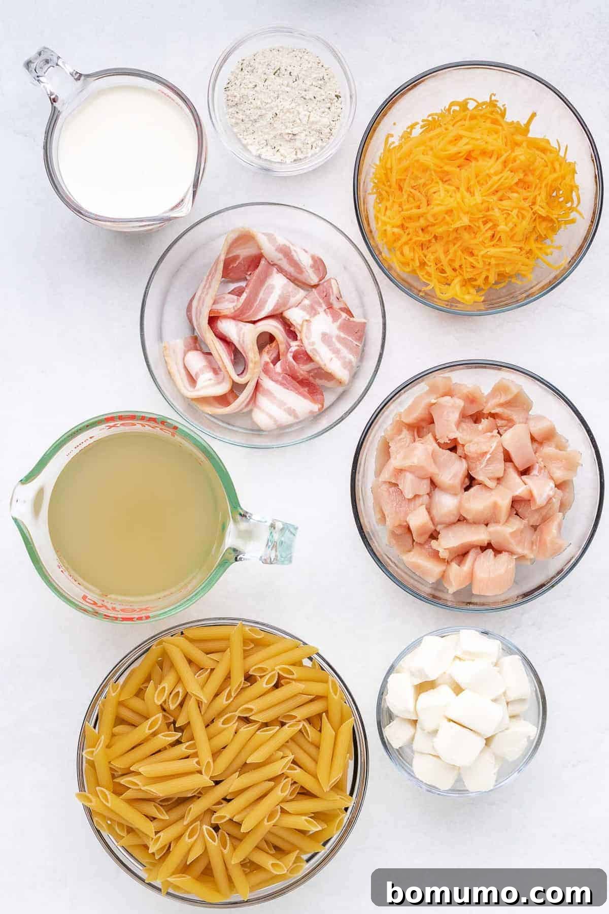 Overhead view of neatly arranged glass bowls containing all the ingredients for Instant Pot Crack Chicken Pasta: uncooked pasta, cubed chicken, crispy bacon, softened cream cheese, heavy cream, ranch seasoning, and chicken broth.