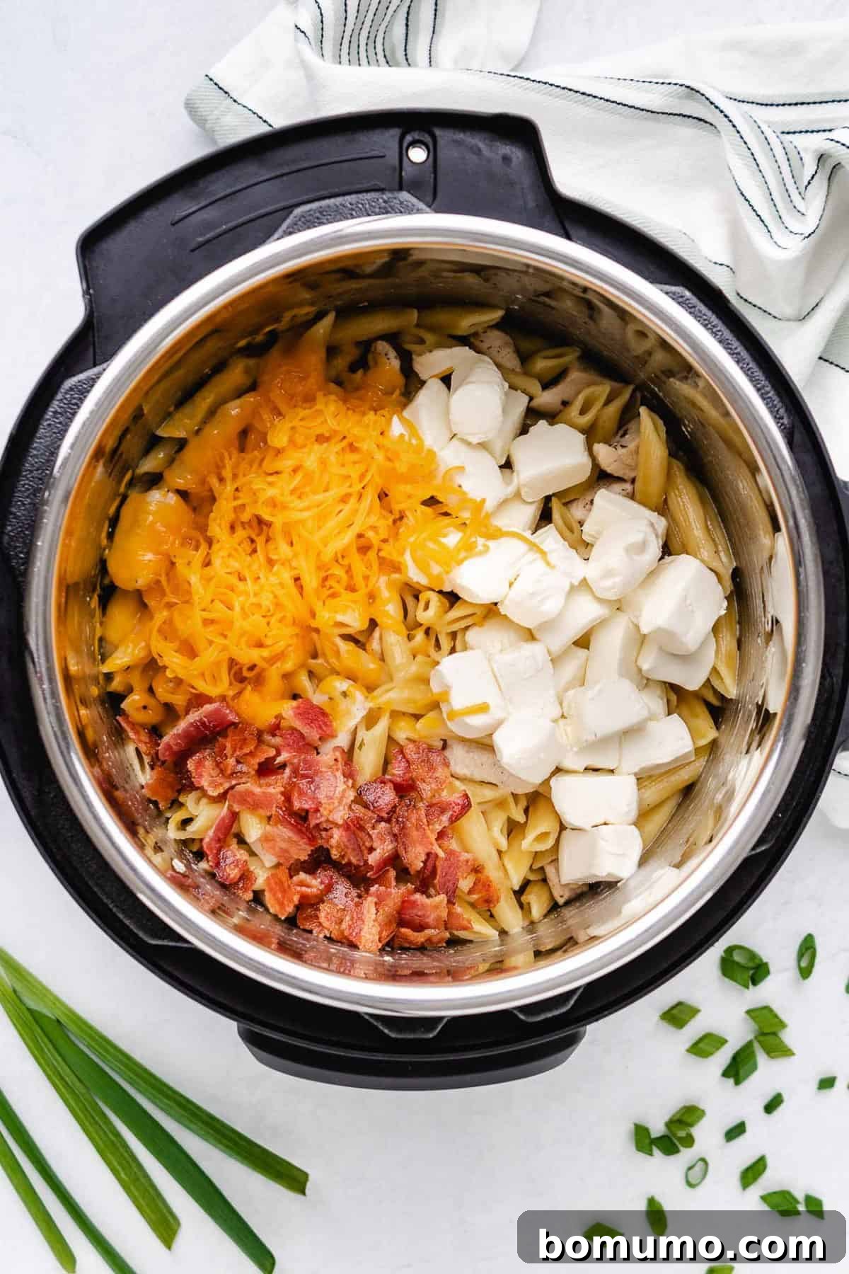 Cheddar cheese and cubed cream cheese melting into the hot cooked pasta in an Instant Pot, with crispy bacon awaiting.