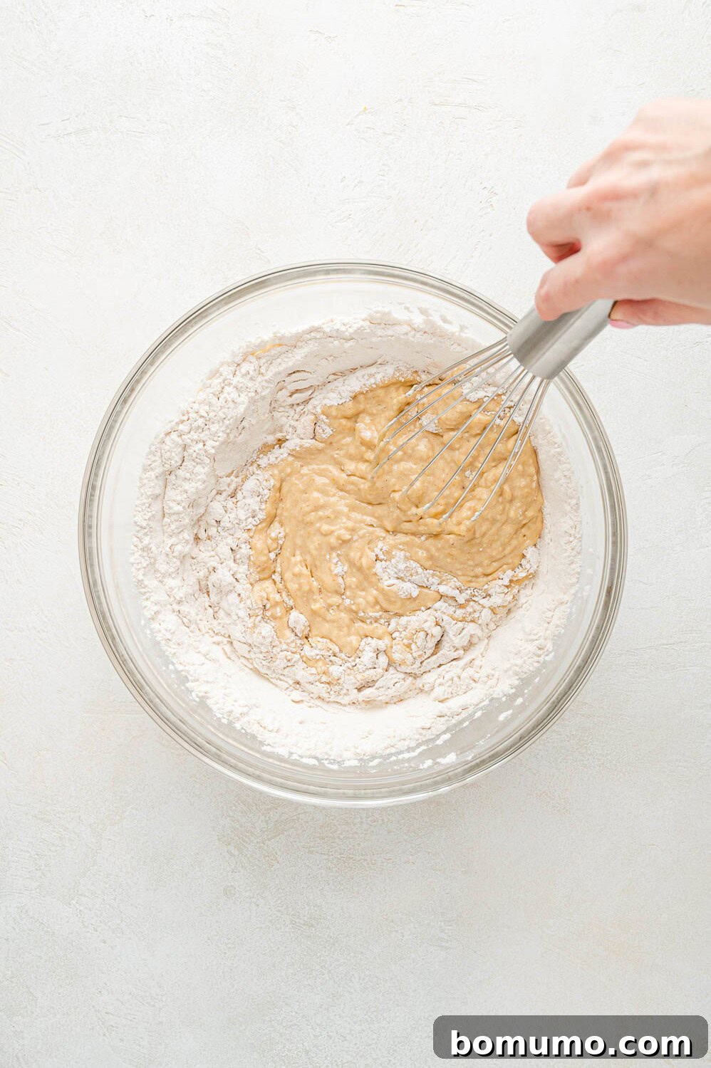 A whisk gently incorporating the dry ingredients into the wet peach pancake batter, aiming for a smooth consistency without overmixing.