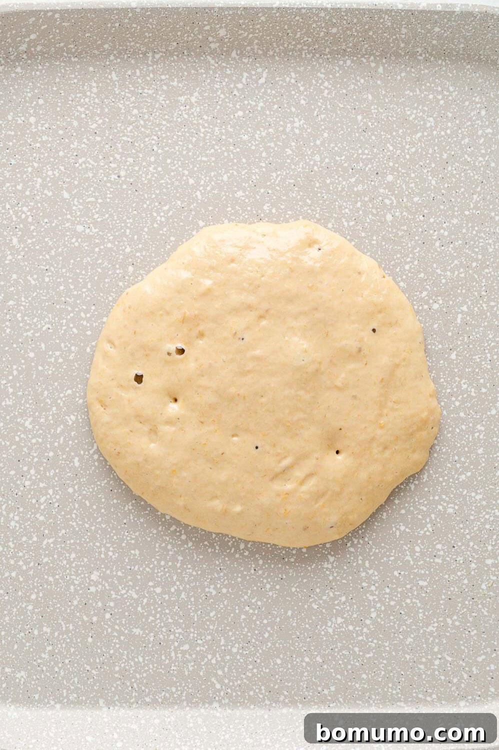 A peach pancake cooking on a griddle, showing bubbles forming on the surface, indicating it's almost ready to flip.