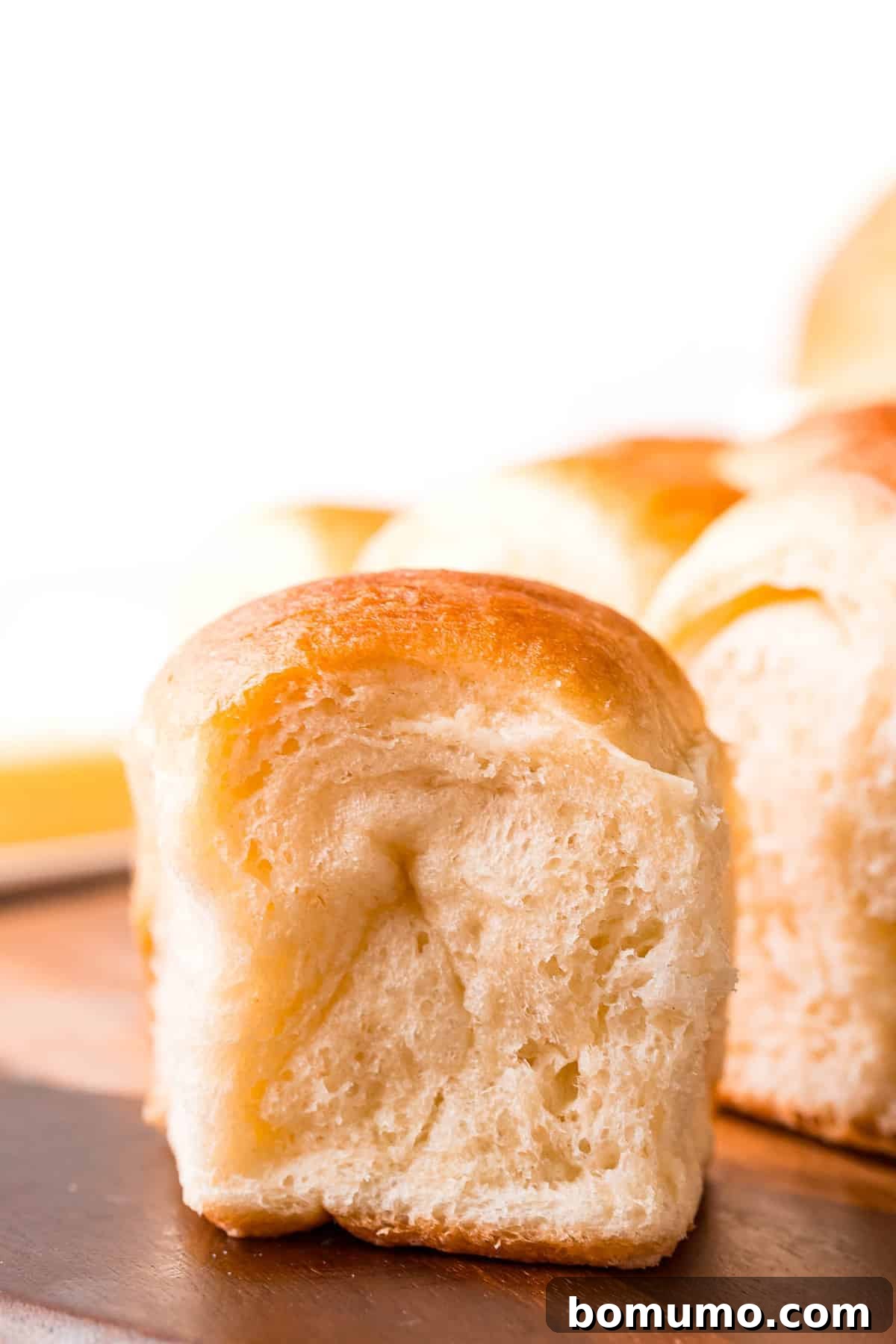 Golden brown Parker House roll on a rustic wooden surface, with more perfectly baked rolls blurred in the background, emphasizing its soft texture.