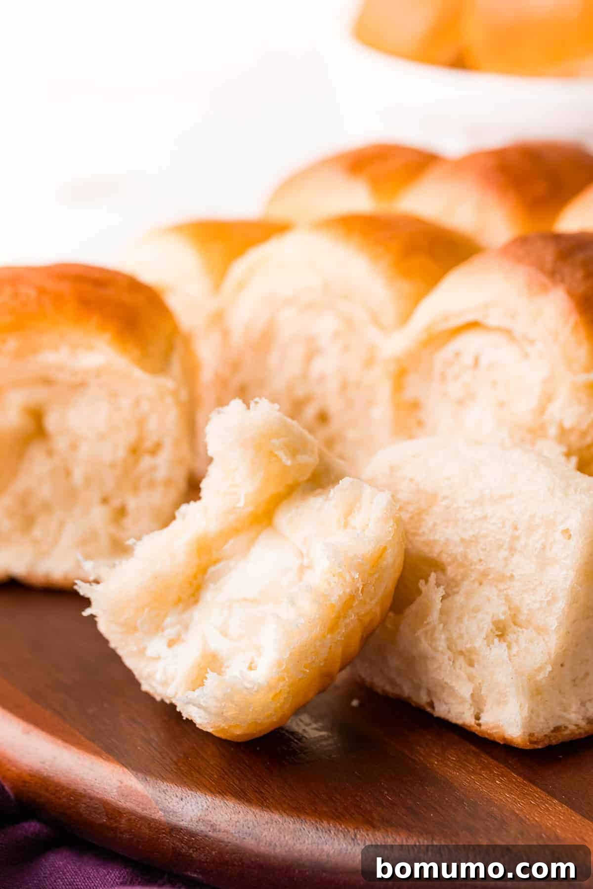 A soft, flaky Parker House roll with a piece gently torn off, revealing its tender, airy interior, with more rolls in the background.