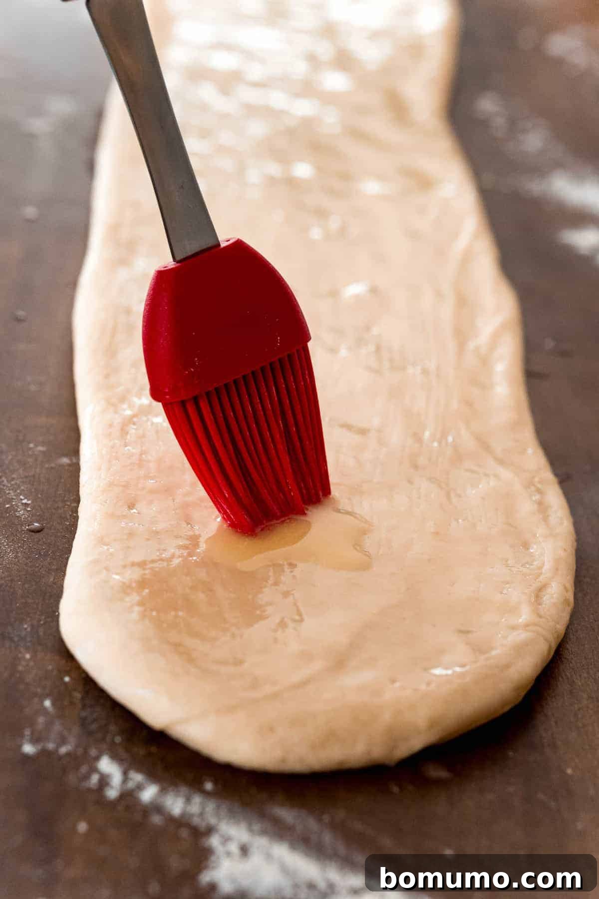 Melted butter being brushed onto a rectangle of yeasted dough, preparing it for the iconic Parker House fold.