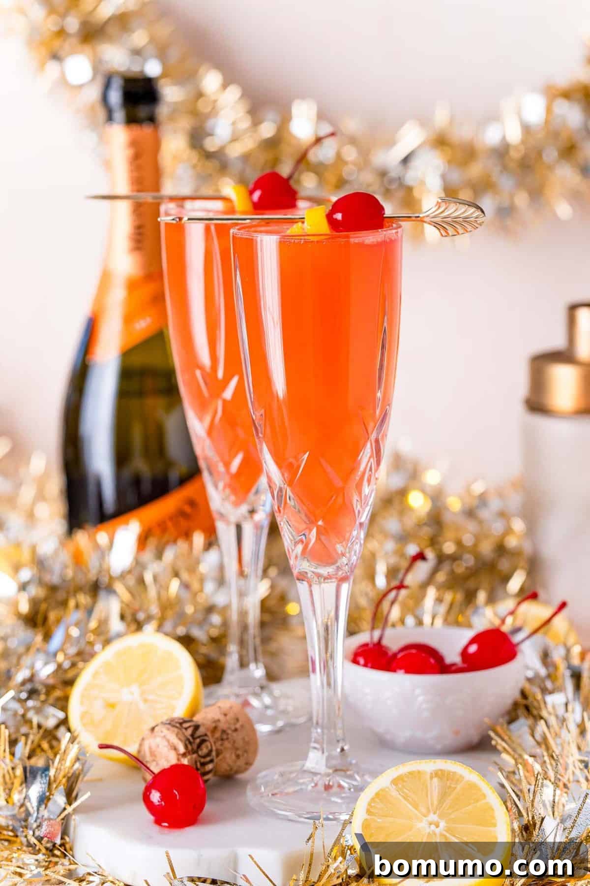 Two champagne flutes with a light red drink garnished with cherries and lemon.