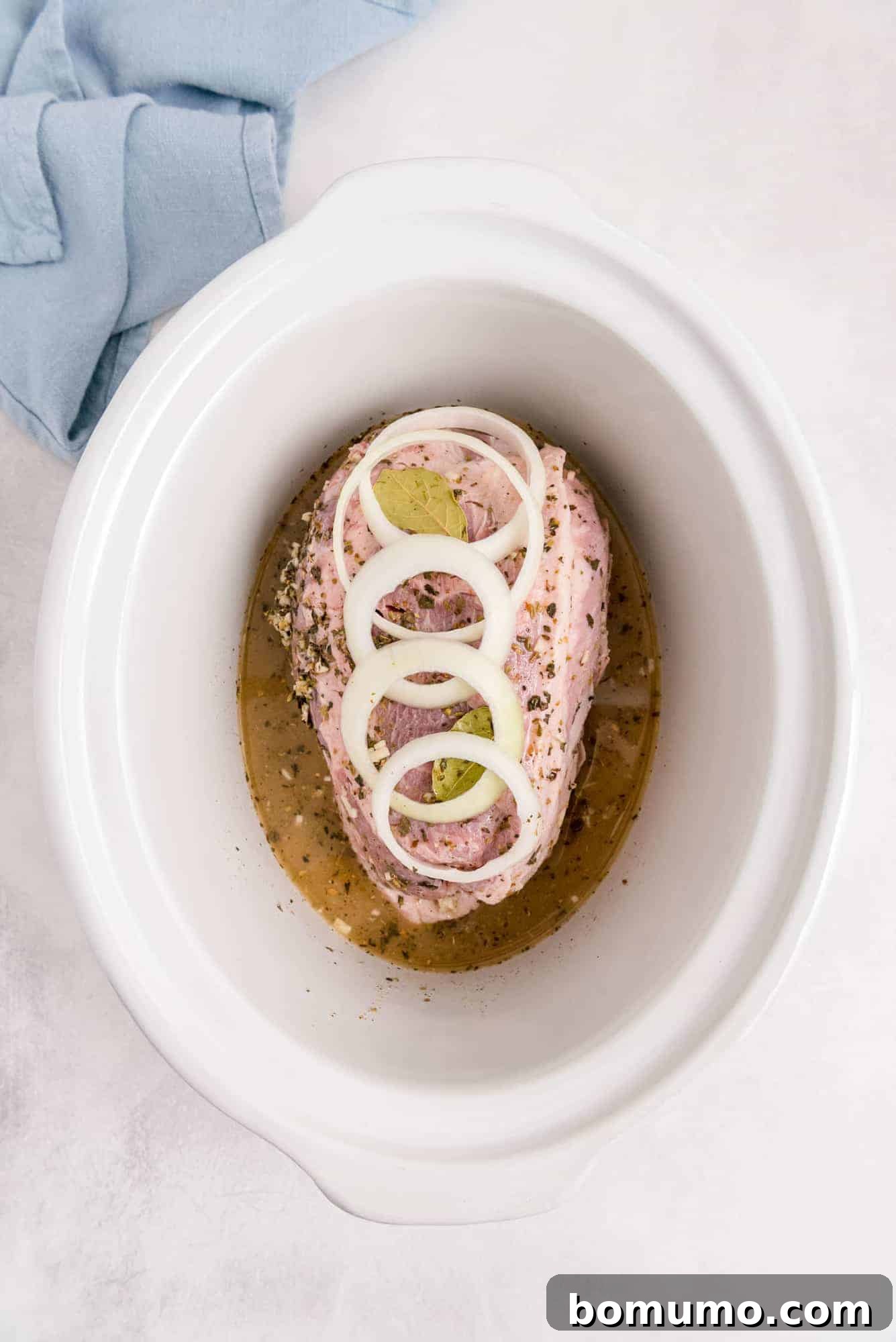 Uncooked pork shoulder with a generous layer of sliced onions and the flavorful marinade in a white slow cooker, ready to begin the slow cooking process.