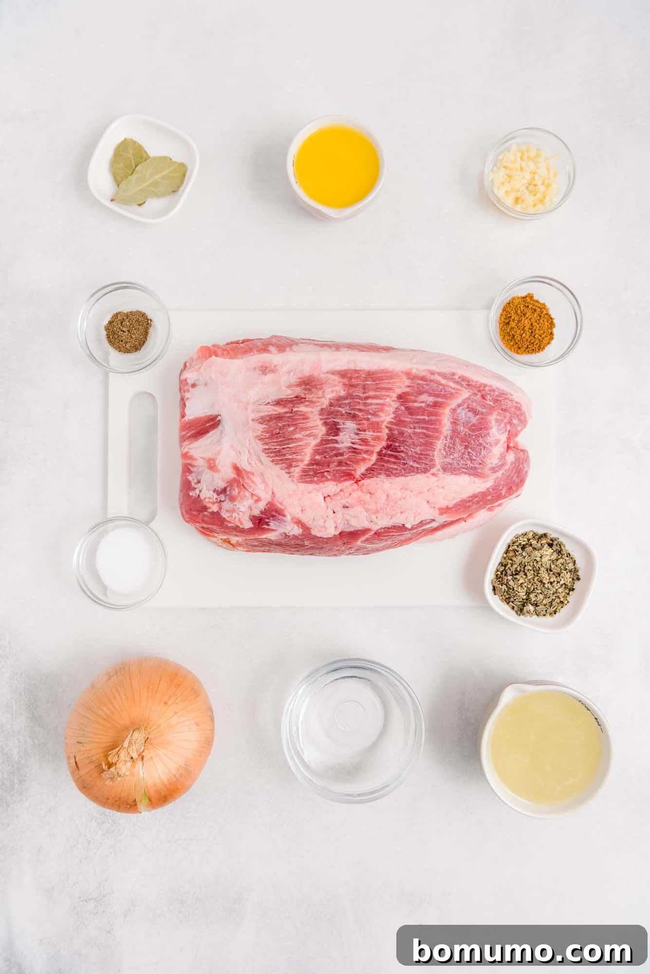 Overhead flat lay of all fresh ingredients for Slow Cooker Cuban Pork, including pork shoulder, a large onion, fresh limes and oranges, and an array of spices.