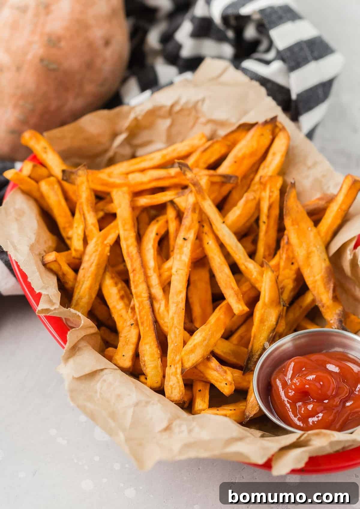 Overhead view of crispy air fryer sweet potato fries served in a rustic brown paper-lined basket, accompanied by a small cup of vibrant red ketchup.