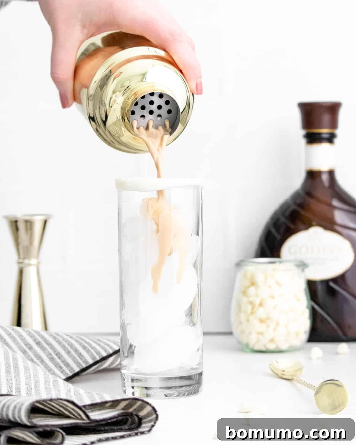 A White Chocolate White Russian cocktail being skillfully poured from a sleek shaker into an elegant glass filled with ice, ready for serving.