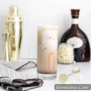 A White Russian in a tall glass, with ingredients and a shaker in the background, ready to be mixed.