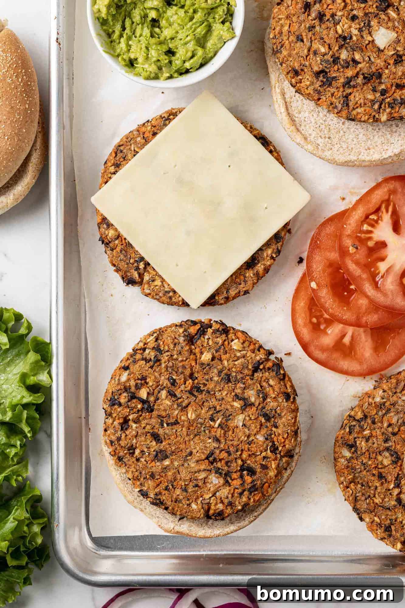 Savory Black Bean Bliss 11 Cooked black bean burgers on a baking sheet, with one patty topped with a slice of melted cheese. Various fresh toppings like lettuce, tomato, and onion are visible nearby, ready for assembly.