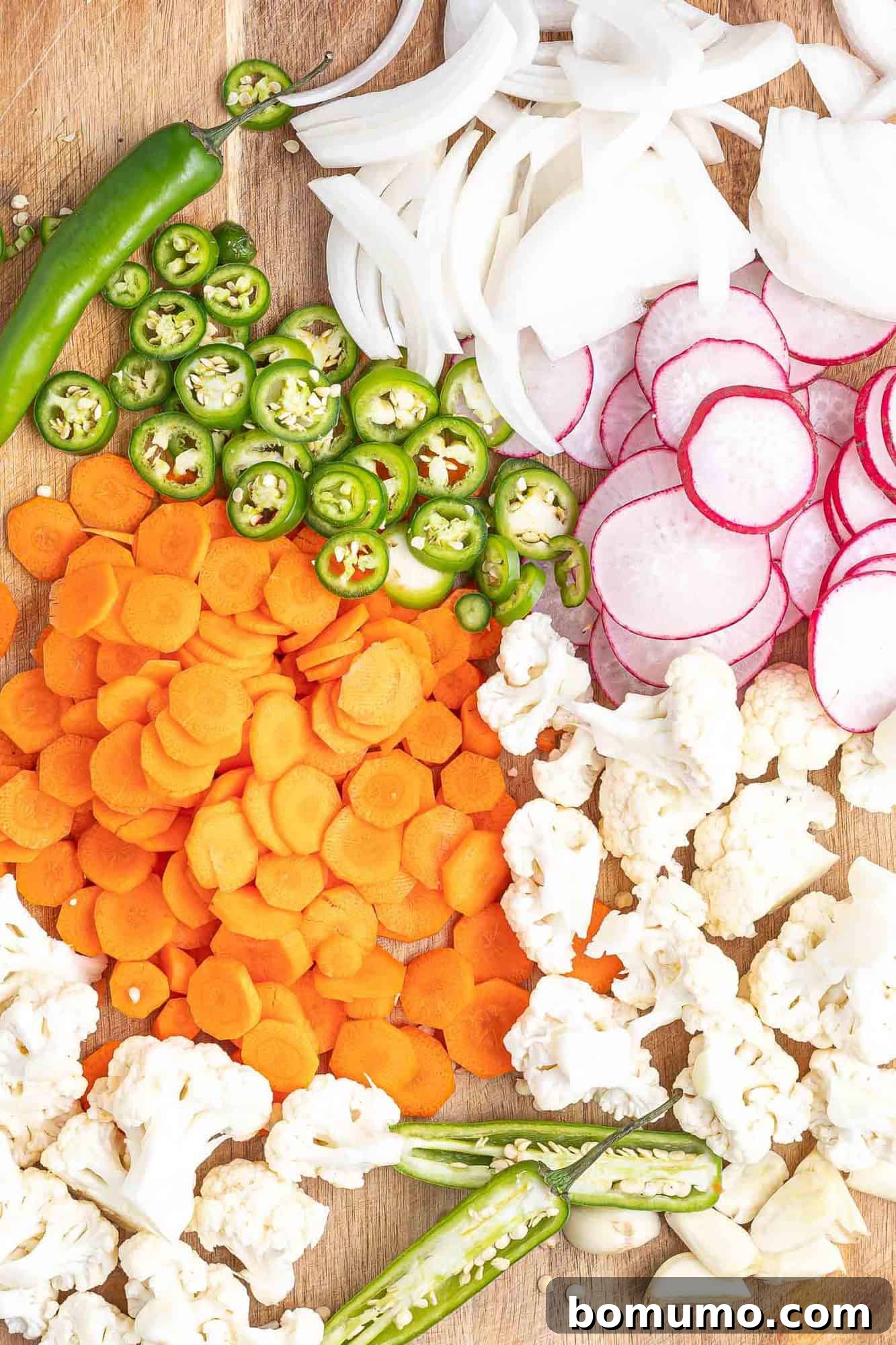Assorted cut vegetables laid out on a wooden surface, including radishes, jalapeño, carrots, cauliflower, and onion.