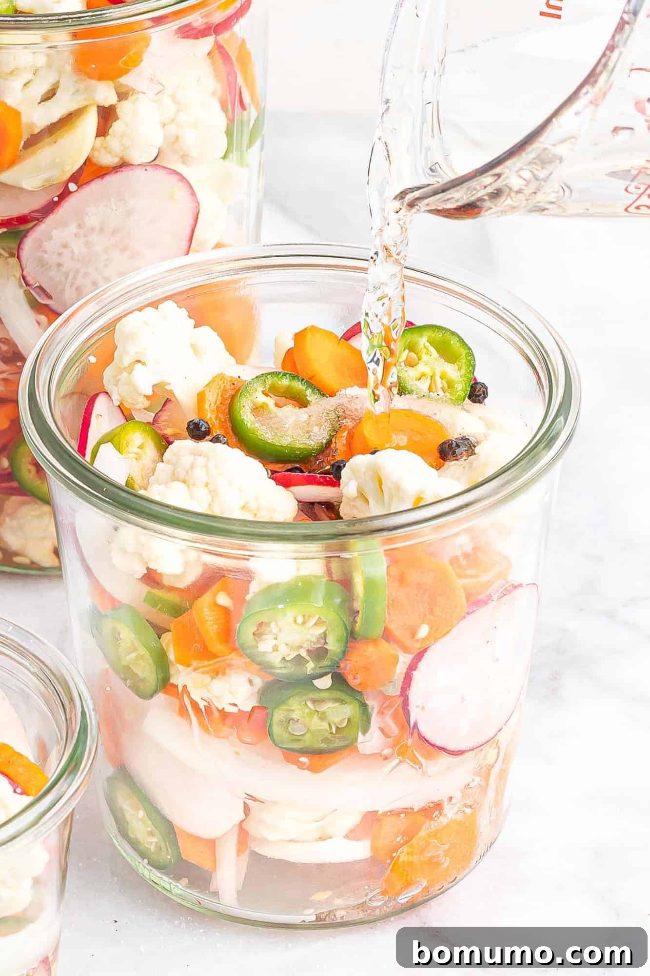 Boiling liquid being carefully poured into a jar filled with cut vegetables, sugar, salt, and peppercorns.