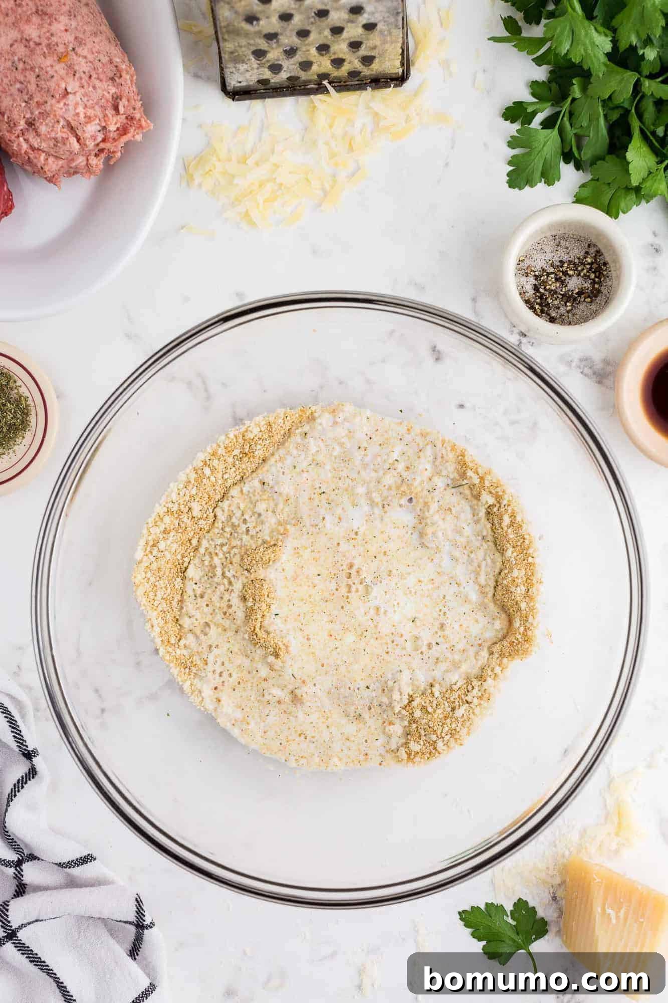 A close-up image of breadcrumbs soaking in milk within a mixing bowl, beginning to soften.
