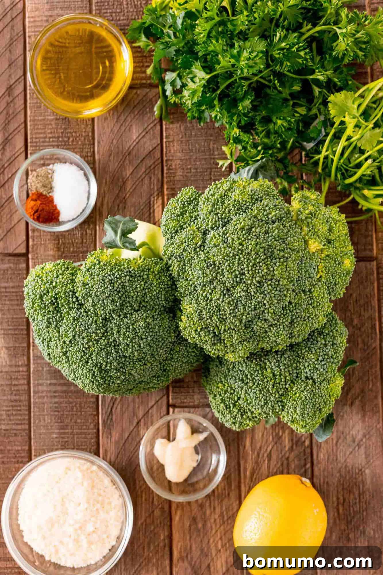 Crispy Lemon-Parmesan Roasted Broccoli 3 Overhead view of fresh broccoli crowns, olive oil, garlic powder, salt, pepper, paprika, a lemon, and grated Parmesan cheese, neatly arranged as ingredients.