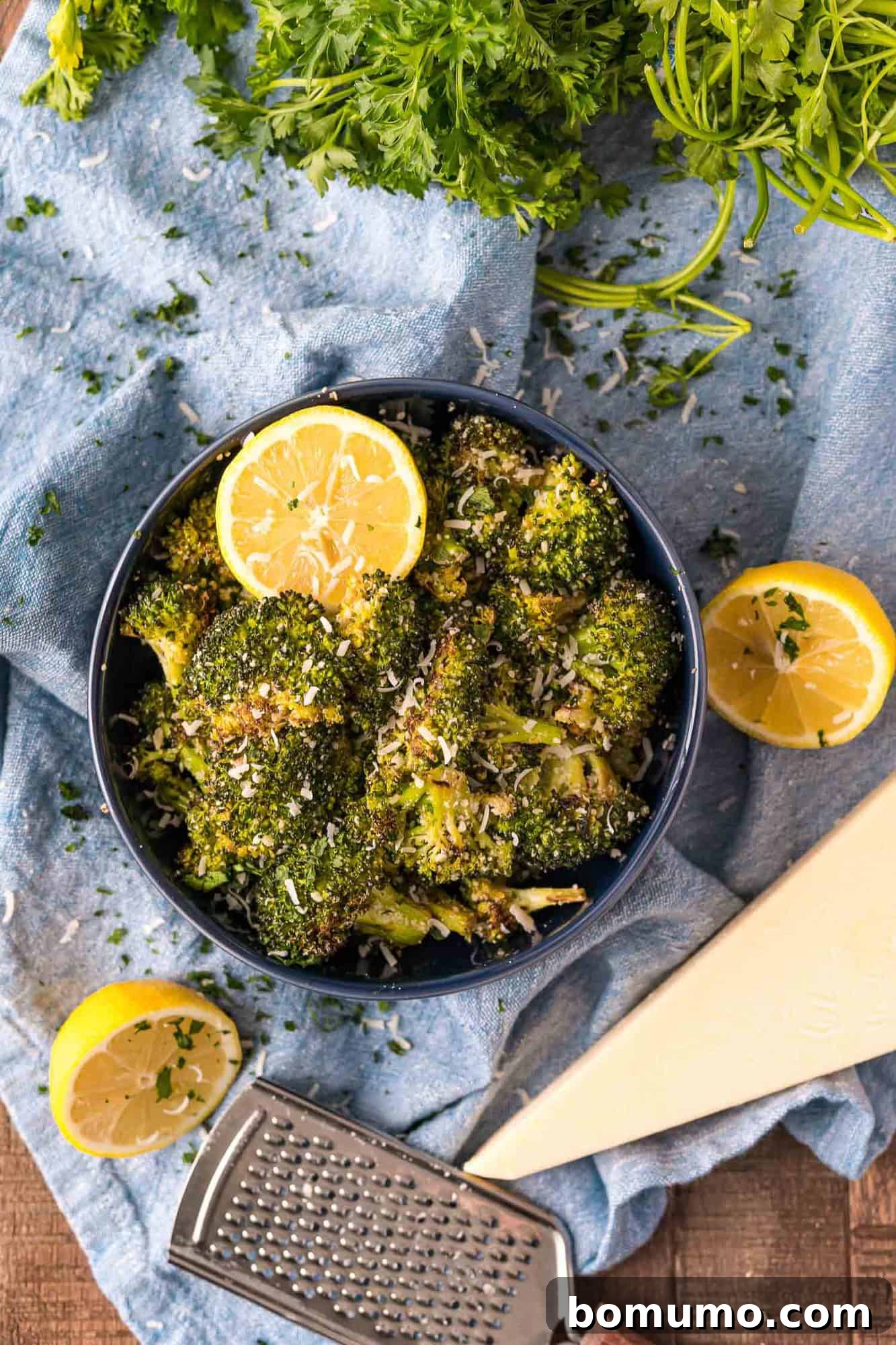 Crispy Lemon-Parmesan Roasted Broccoli 9 A dark bowl of vibrant roasted broccoli, artfully garnished with fresh lemon wedges, grated Parmesan, and chopped parsley, symbolizing freshness and flavor.