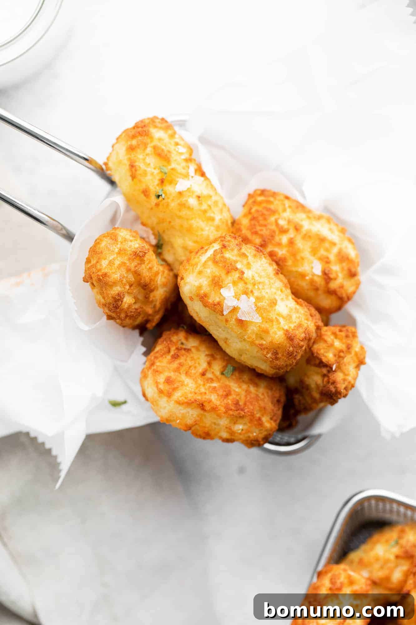 Overhead view of crispy air fryer tater tots in a small metal basket on a white background, invitingly golden brown.