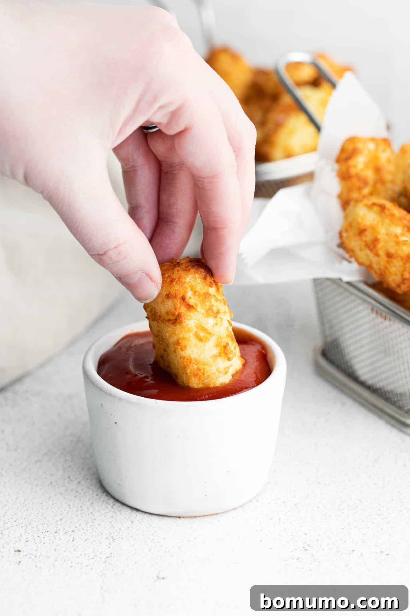 A perfectly cooked homemade tater tot being dipped into a small white dish of bright red ketchup, highlighting its crispy texture.