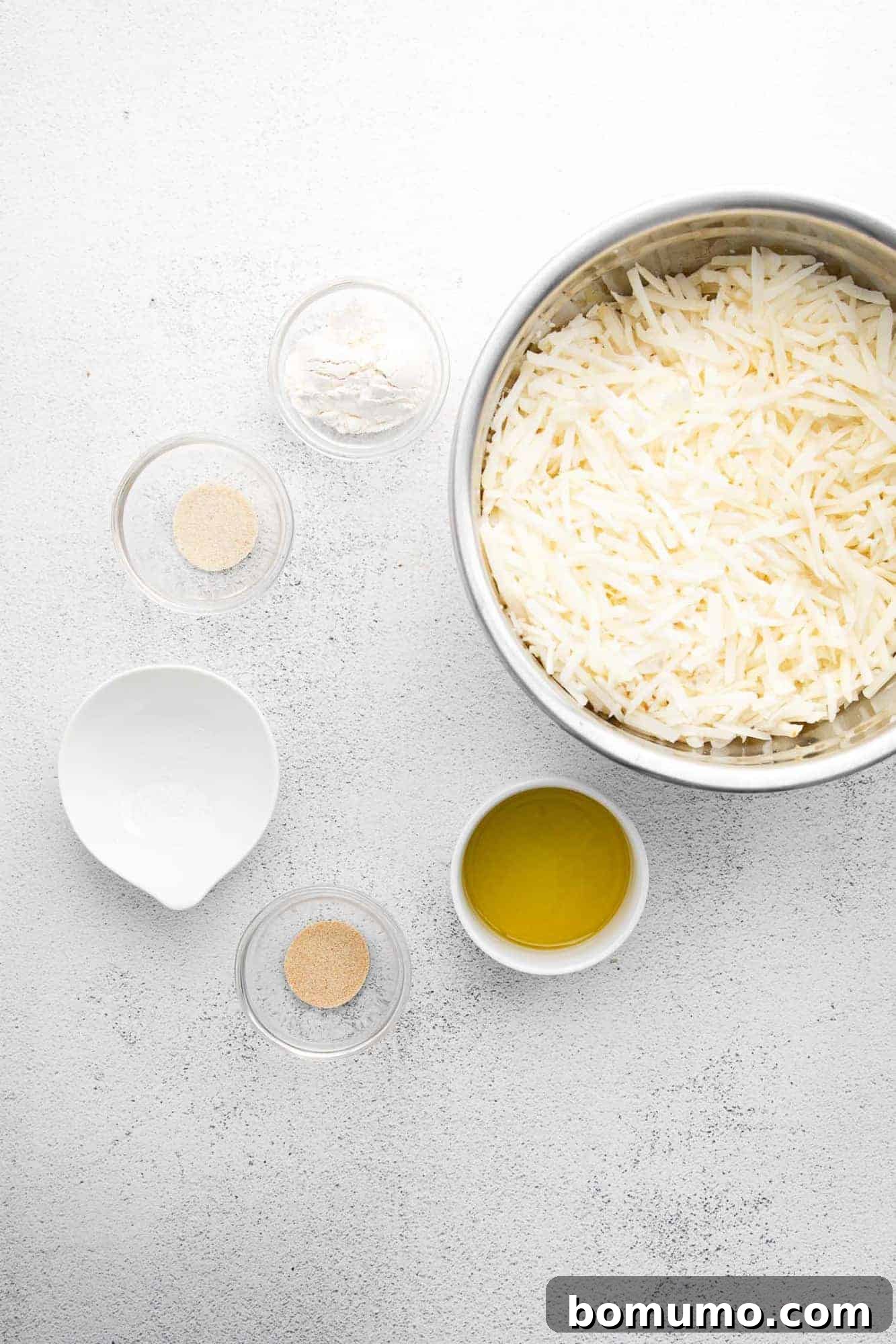 Overhead view of fresh ingredients neatly arranged: a bowl of shredded potatoes, a bottle of olive oil, a bowl of white flour, and small dishes of various spices, ready for mixing.