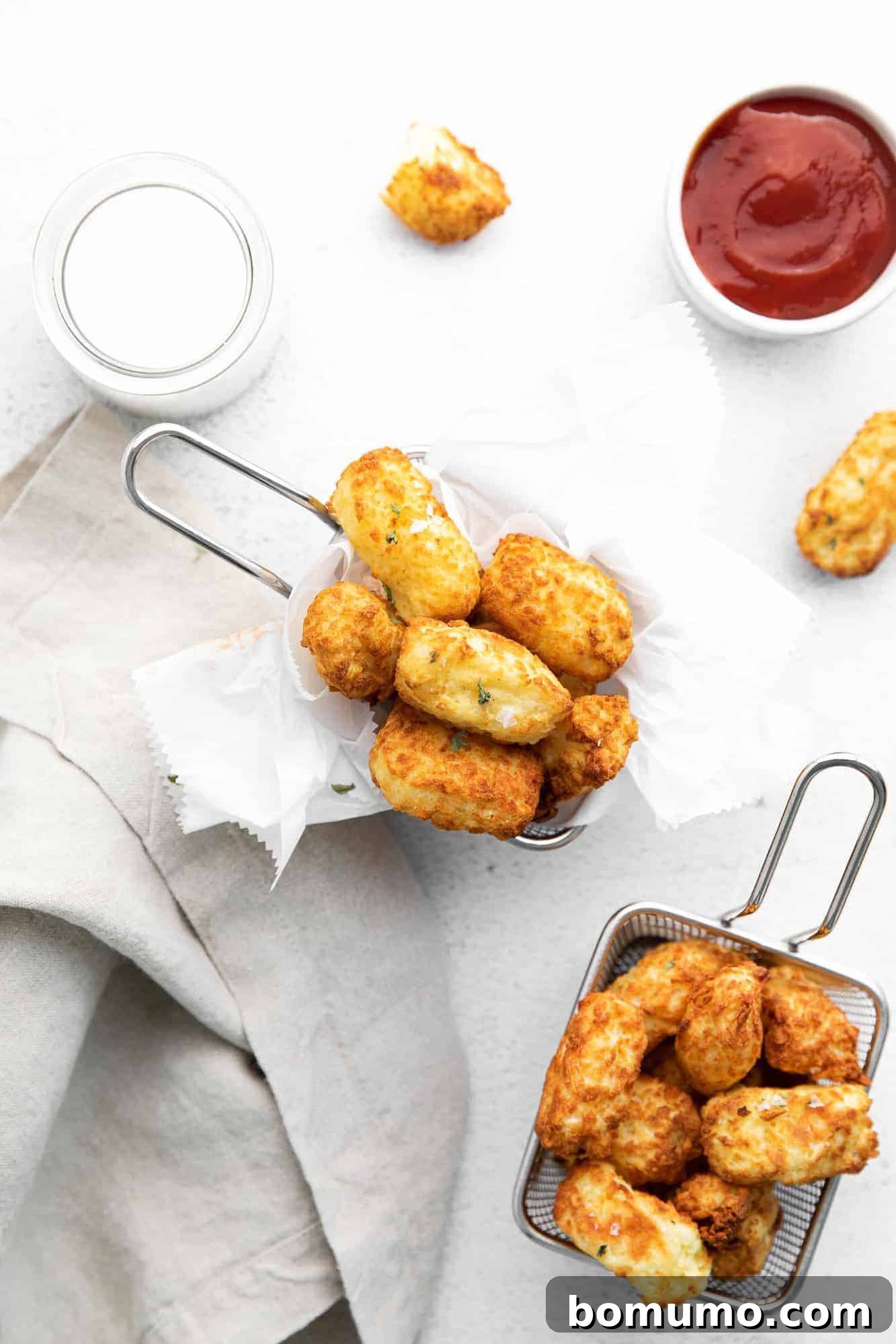 Overhead view of small metal baskets overflowing with crispy air fryer tater tots, accompanied by a small white dish of ketchup for dipping.