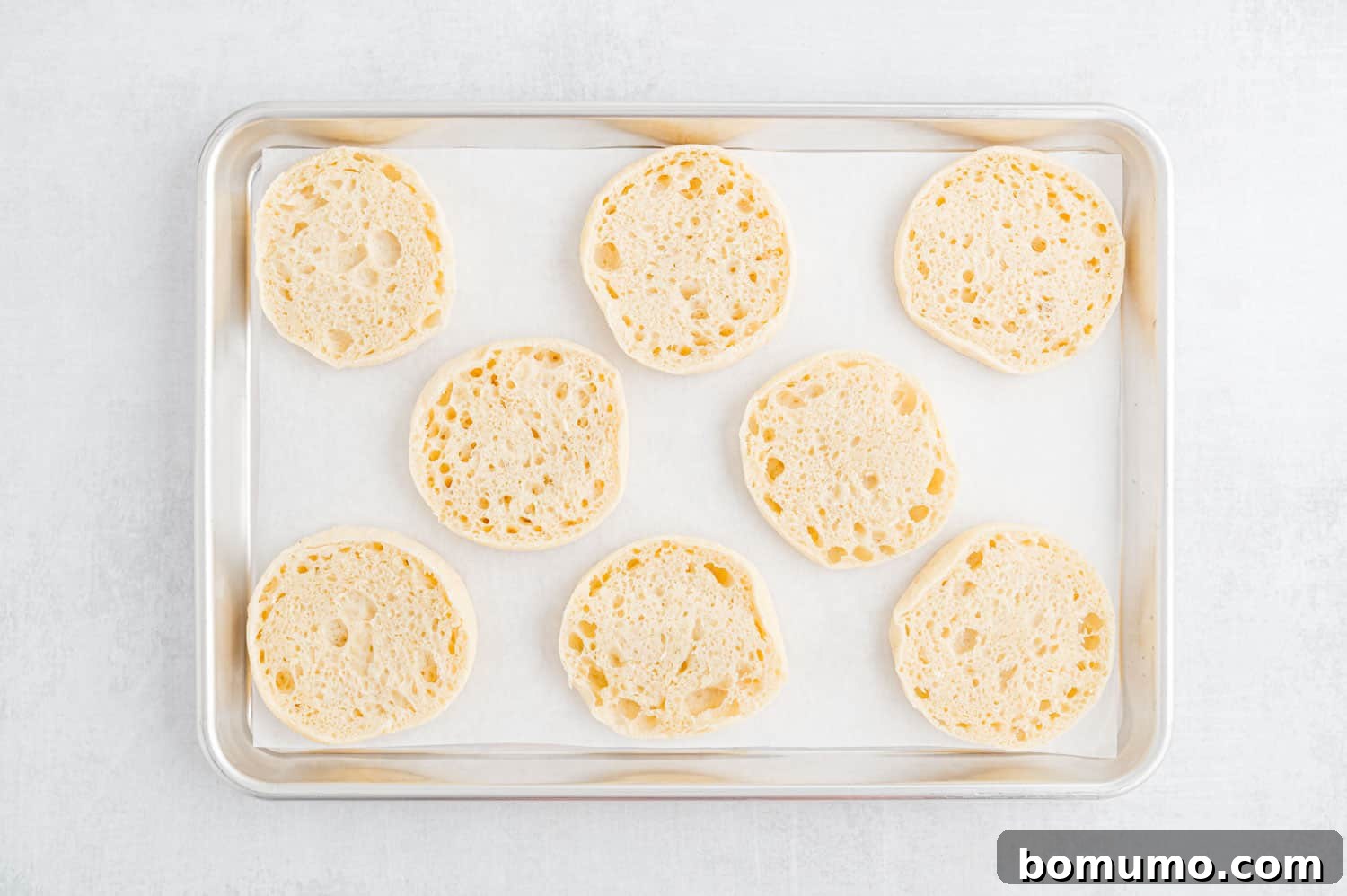 English muffins cut in half, revealing their characteristic nooks and crannies, arranged on a sheet pan lined with parchment paper.