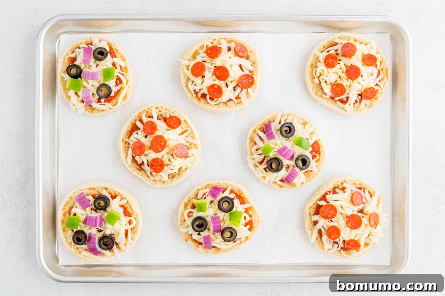 Uncooked English muffin pizzas, topped with sauce, cheese, and pepperoni, neatly arranged on a parchment paper-lined sheet pan, ready for their final bake.