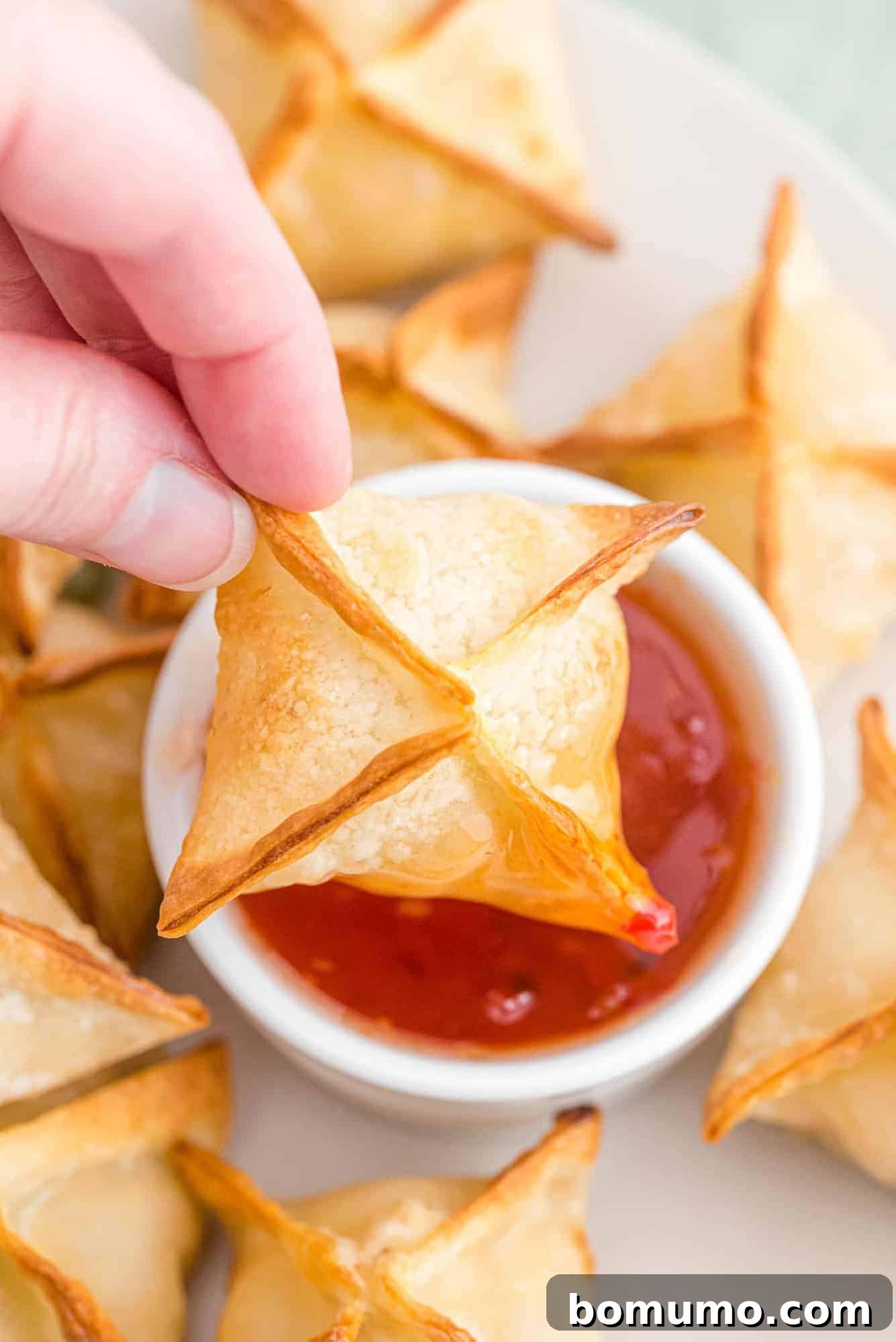 A crispy crab wonton being dipped into a small bowl of glistening sweet chili sauce.