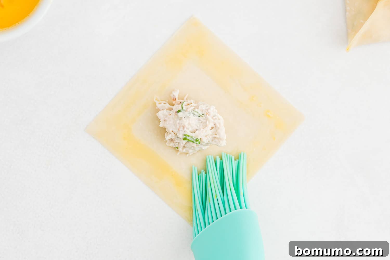 A wonton wrapper being brushed with egg wash on all four edges before folding.
