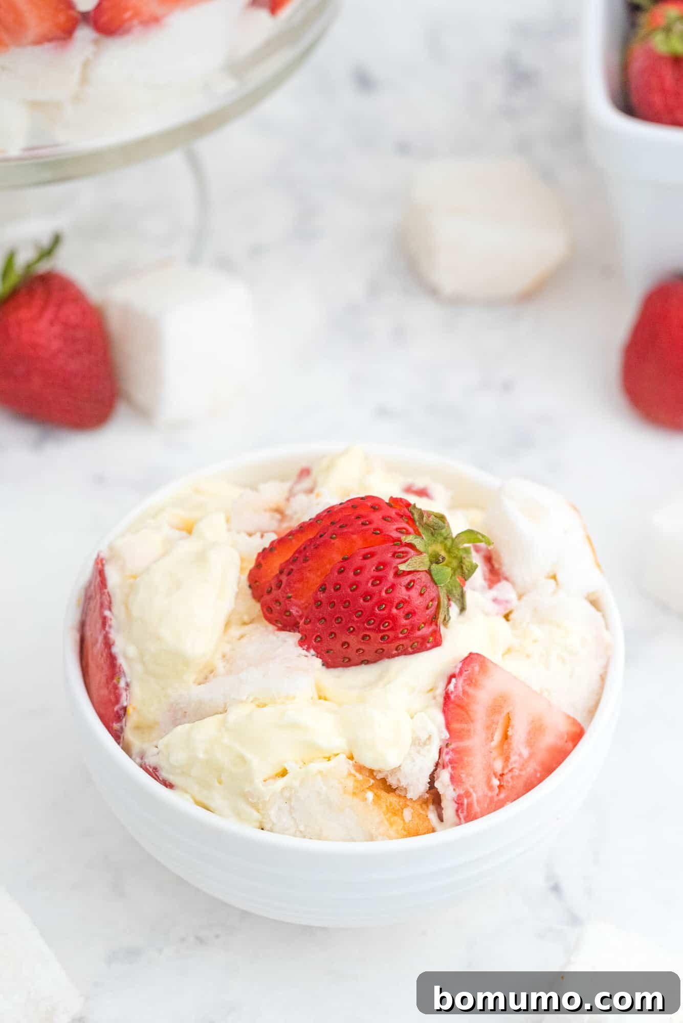 Effortless Strawberry Trifle 13 A portion of strawberry trifle elegantly scooped into a small white bowl and garnished with a fresh strawberry slice.