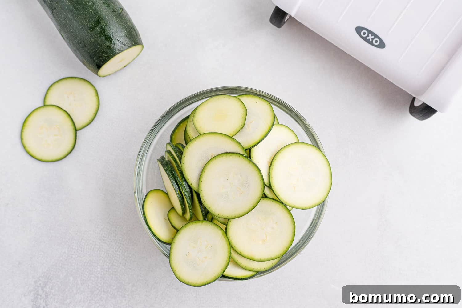 Sliced zucchini in a bowl, a whole zucchini partially visible, with a mandoline slicer in the upper right-hand corner, illustrating the slicing process.