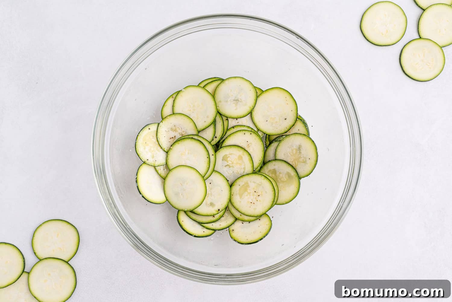 Zucchini slices coated in oil and seasonings, resting in a large glass bowl on a white background.