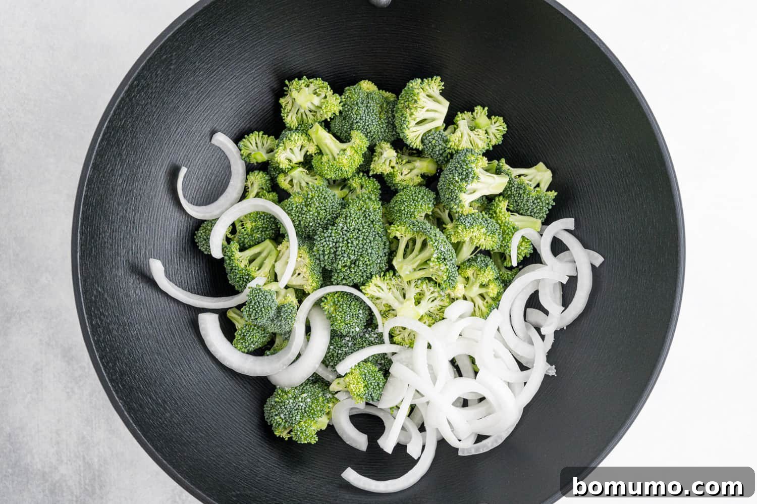 Fresh broccoli florets and sliced white onion stir-frying in a hot black wok, glistening with oil.