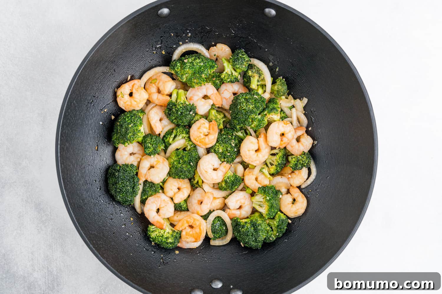 Shrimp, broccoli, and onions coated in a rich brown sauce, all combined in a black wok and actively stir-frying.
