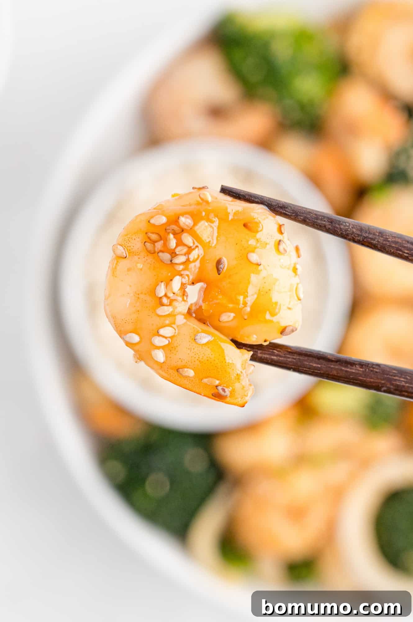 A single piece of shrimp, coated in brown sauce and sesame seeds, held between chopsticks, highlighting the delicious result.
