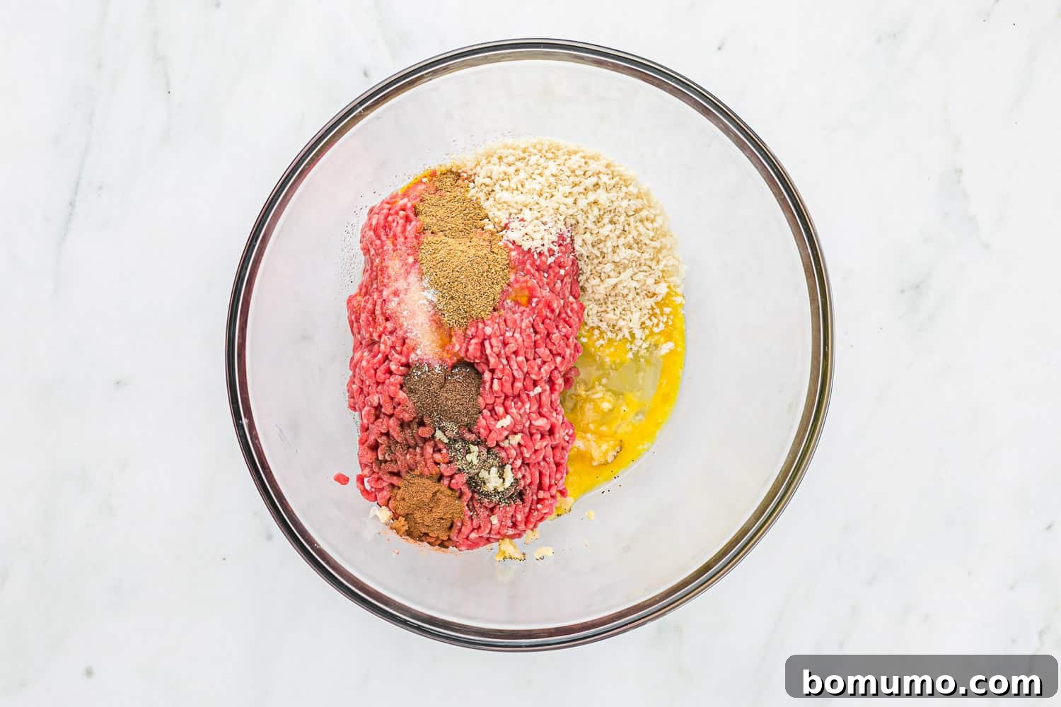 Meatball ingredients in a clear glass mixing bowl, ready to be combined.