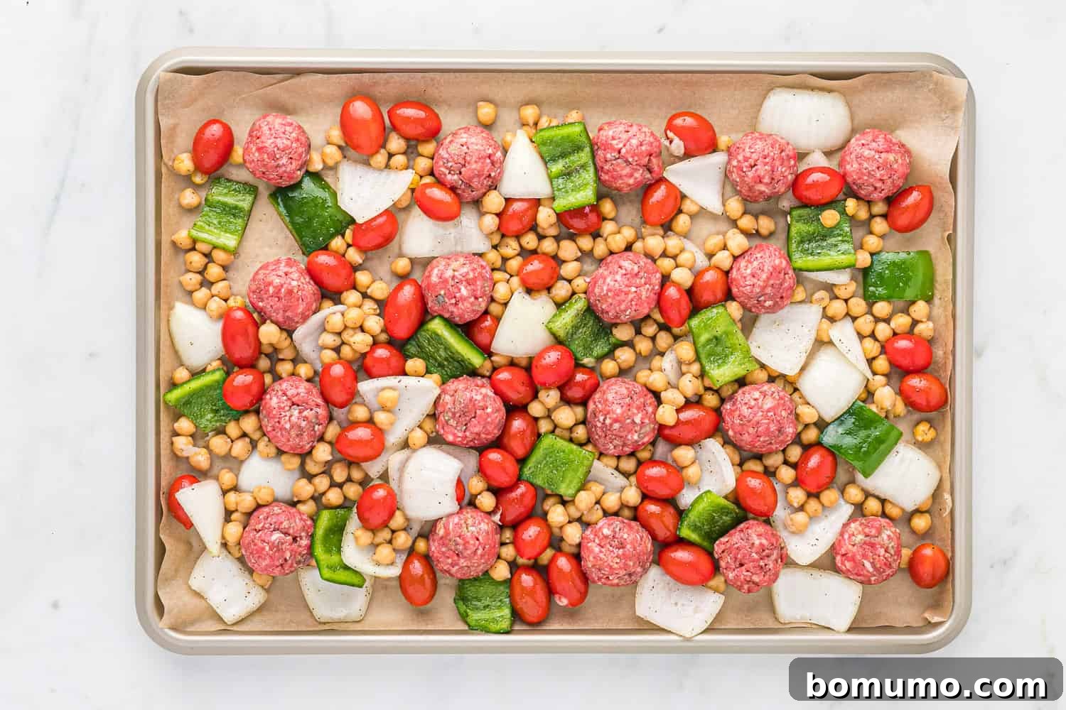 An uncooked sheet pan dinner, with meatballs, vegetables, and chickpeas arranged.