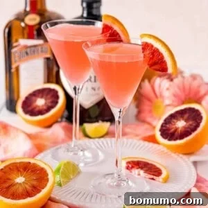 Two light pinkish red cocktails garnished with blood oranges, on a tray.
