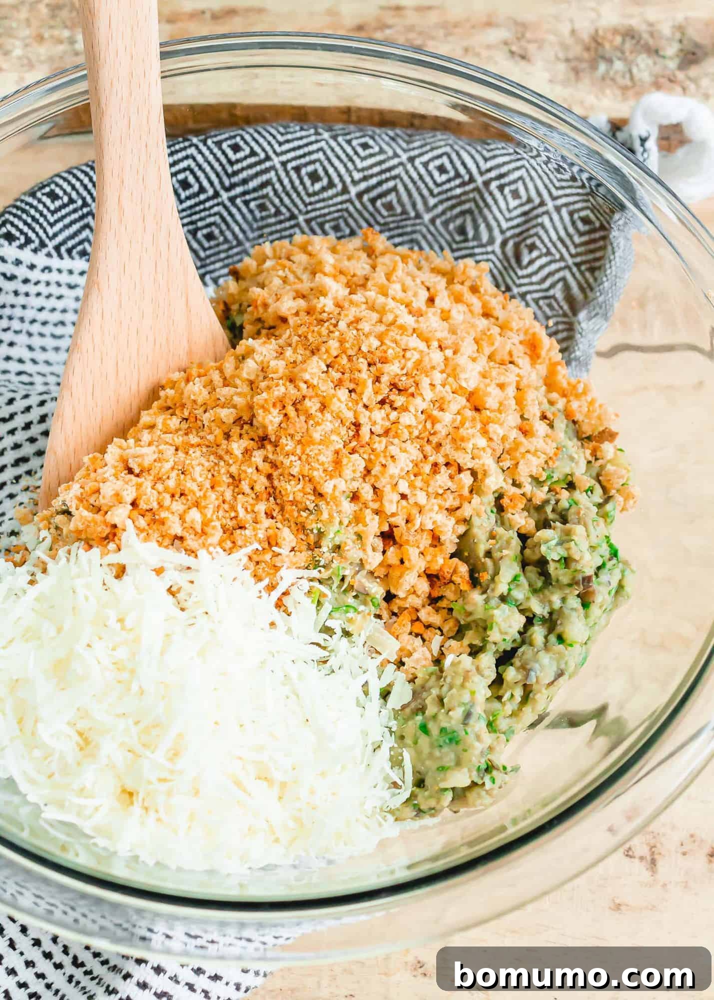 Breadcrumbs and cheese in a clear glass bowl with a wooden spoon.
