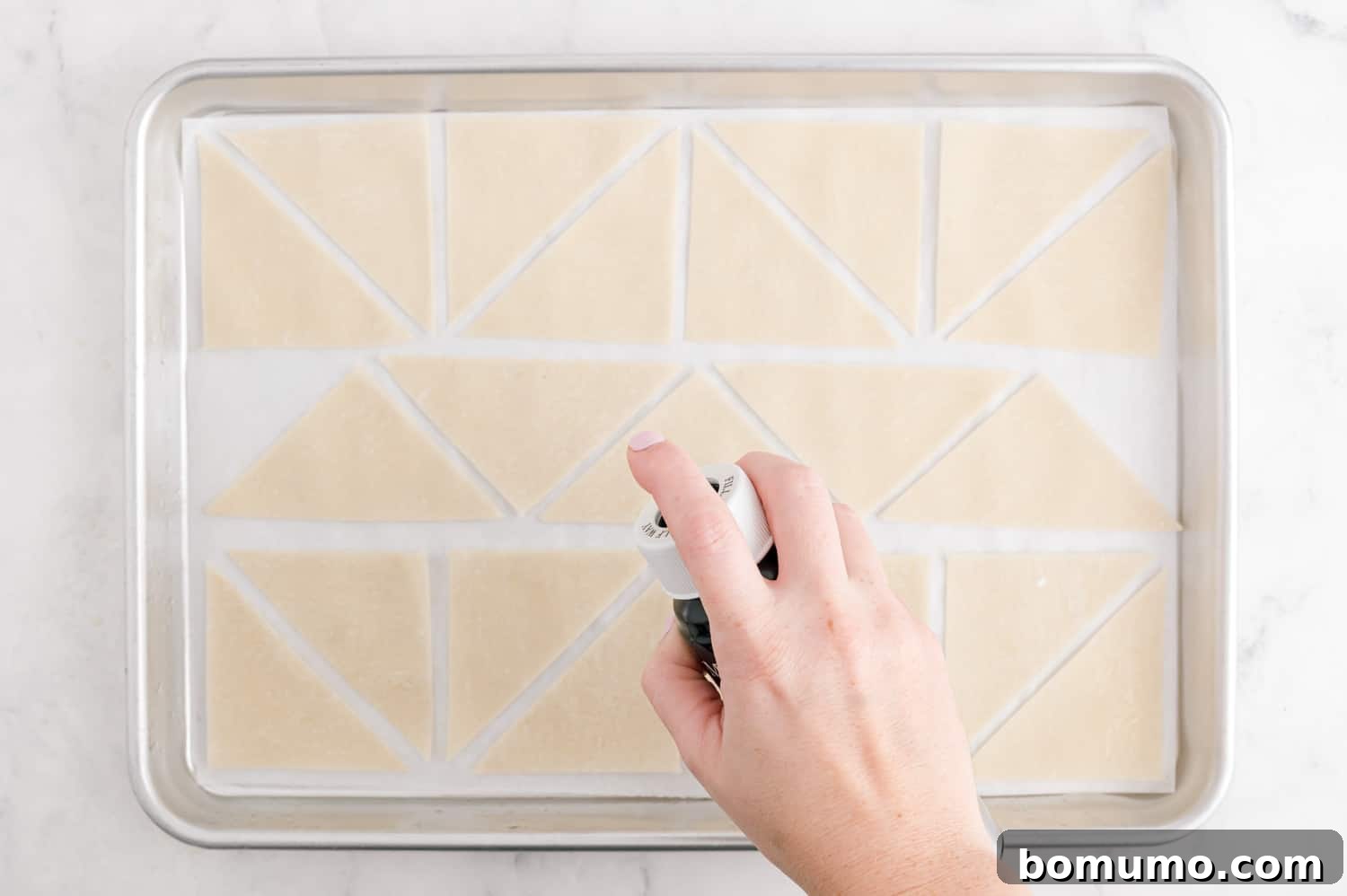 Wonton wrappers being sprayed with oil on a sheet pan.