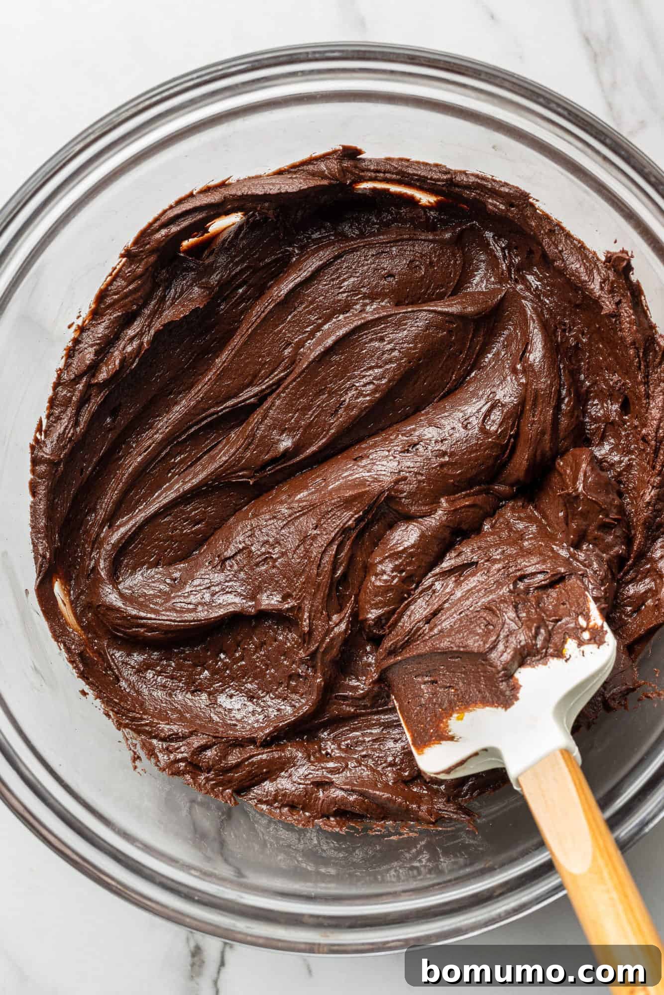 Smooth chocolate frosting in a mixing bowl with a rubber spatula, showcasing its perfect texture.