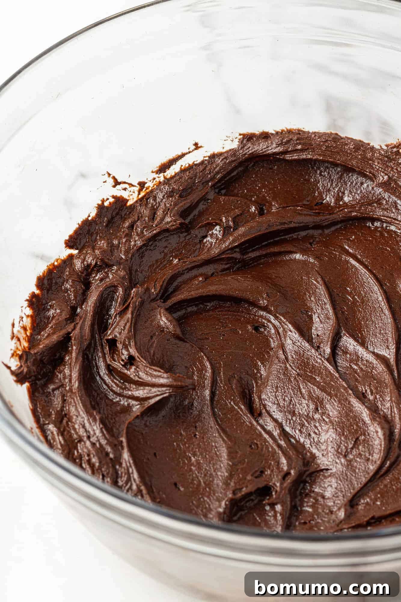 Chocolate frosting in a clear glass mixing bowl, showcasing its smooth, glossy texture after being whipped.