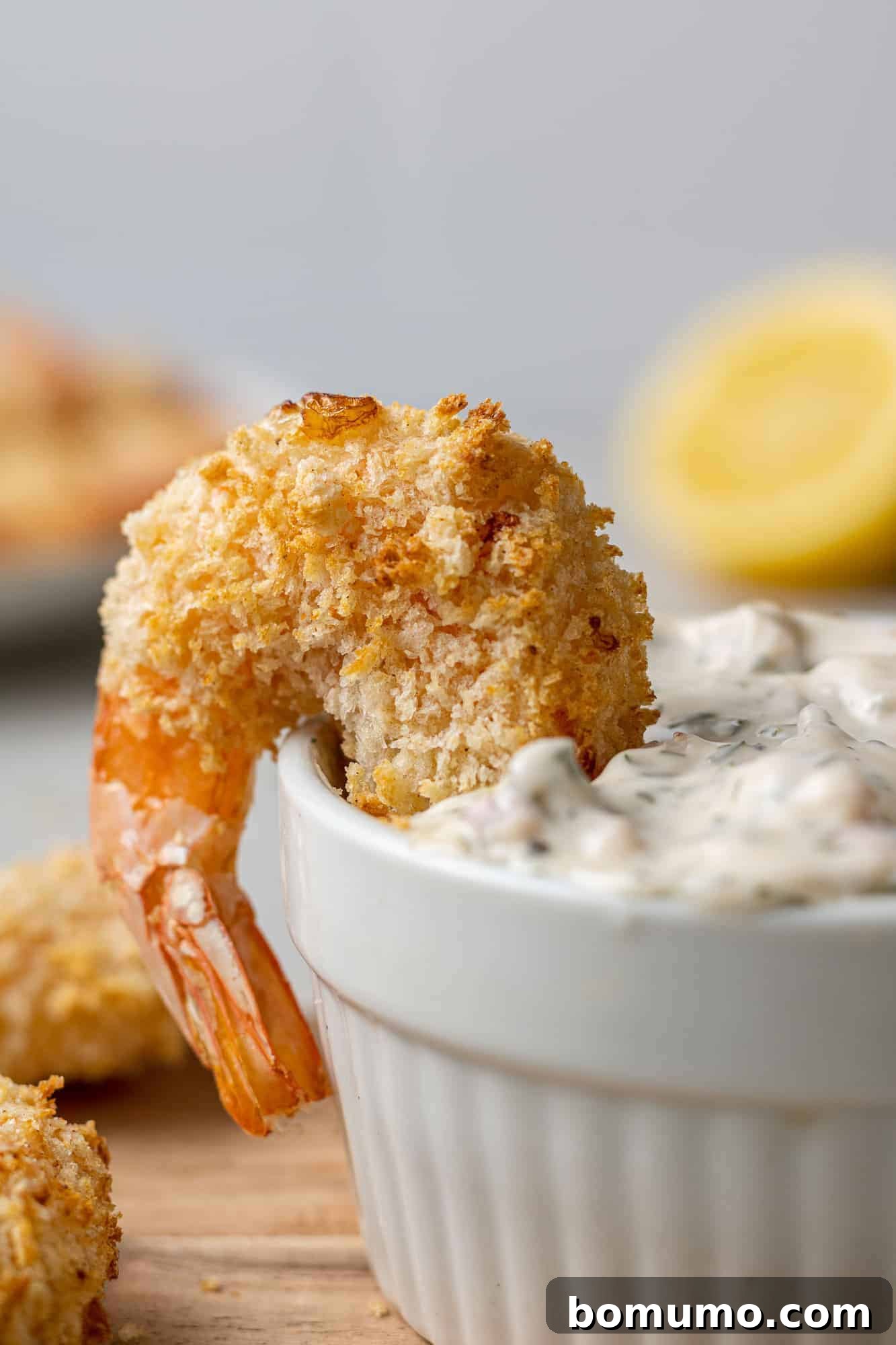 A single piece of crispy air fryer breaded shrimp resting tantalizingly on the rim of a small bowl filled with creamy tartar sauce, perfectly ready for dipping.