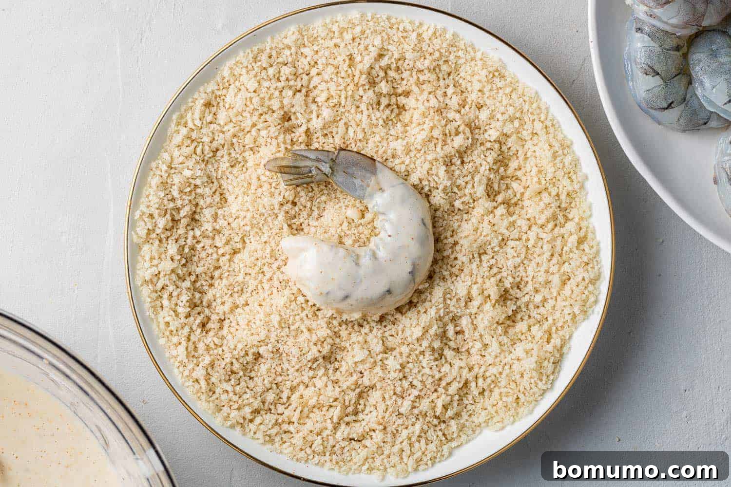 A hand gently rolling a battered shrimp in a bowl of Panko bread crumbs, ensuring a complete and even coating.