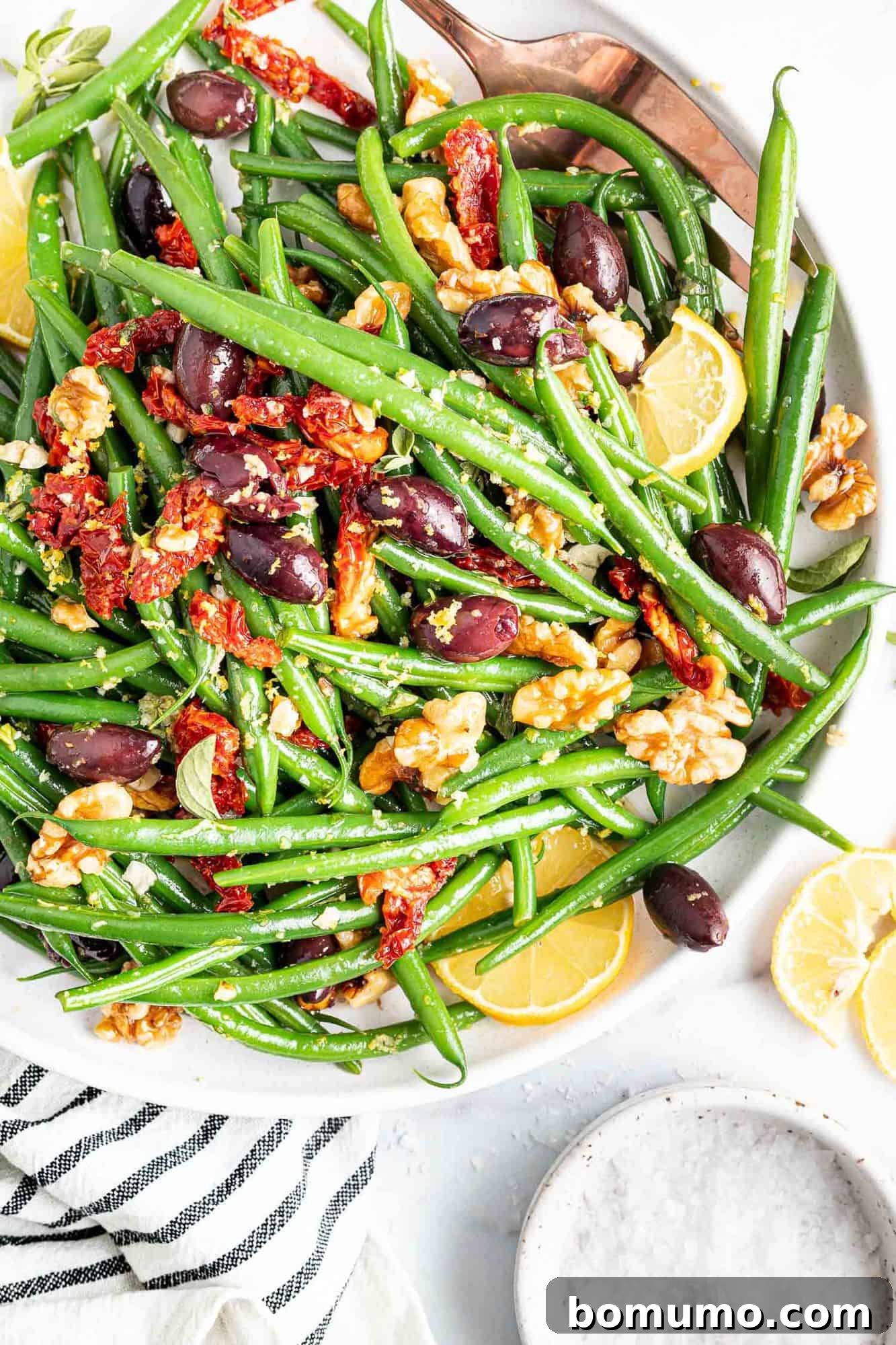 Green beans in a large white bowl with sun dried tomatoes, kalamata olives, and walnuts.