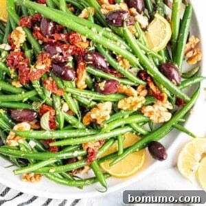 Green beans in a large white bowl with sun dried tomatoes, kalamata olives, and walnuts.